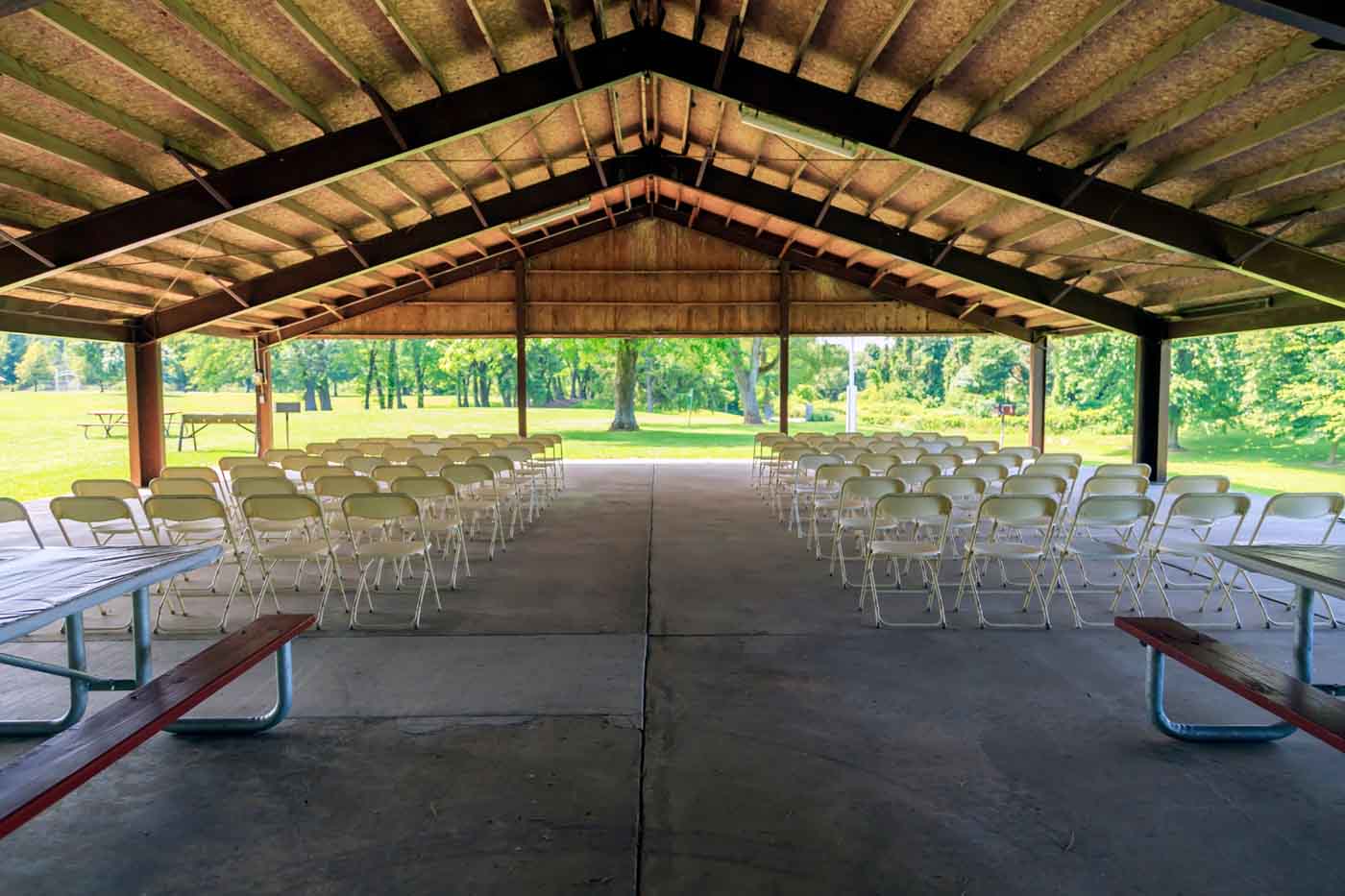 A picnic shelter set up for a wedding ceremony with many rows of white folding chairs. Shot from behind.